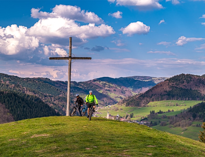 Cycle tour in the Münstertal – mountain biker at a summit cross with a view over the green valley